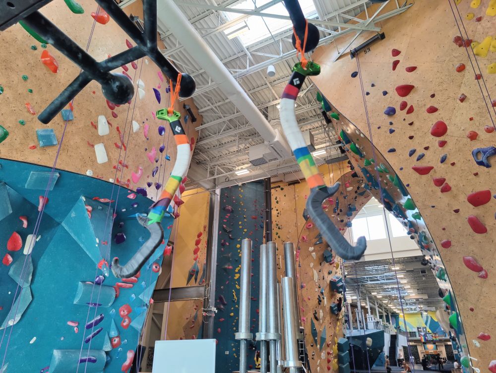 A photo from inside a climbing gym of a pair of the handles for ice tools, with their picks removed, hanging from a pullup bar. The handles are wrapped in tape, which is common for dry tooling (for climbing directly on rock with tools, or competition-style ice climbing)