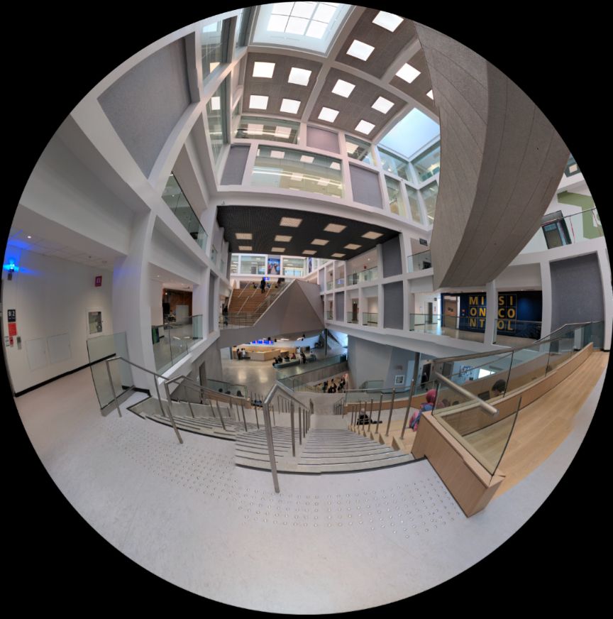 Spherical photo of the interior of a university building. It shows three stories of the building connected through a mix-use wooden staircase and skylights at the top. 