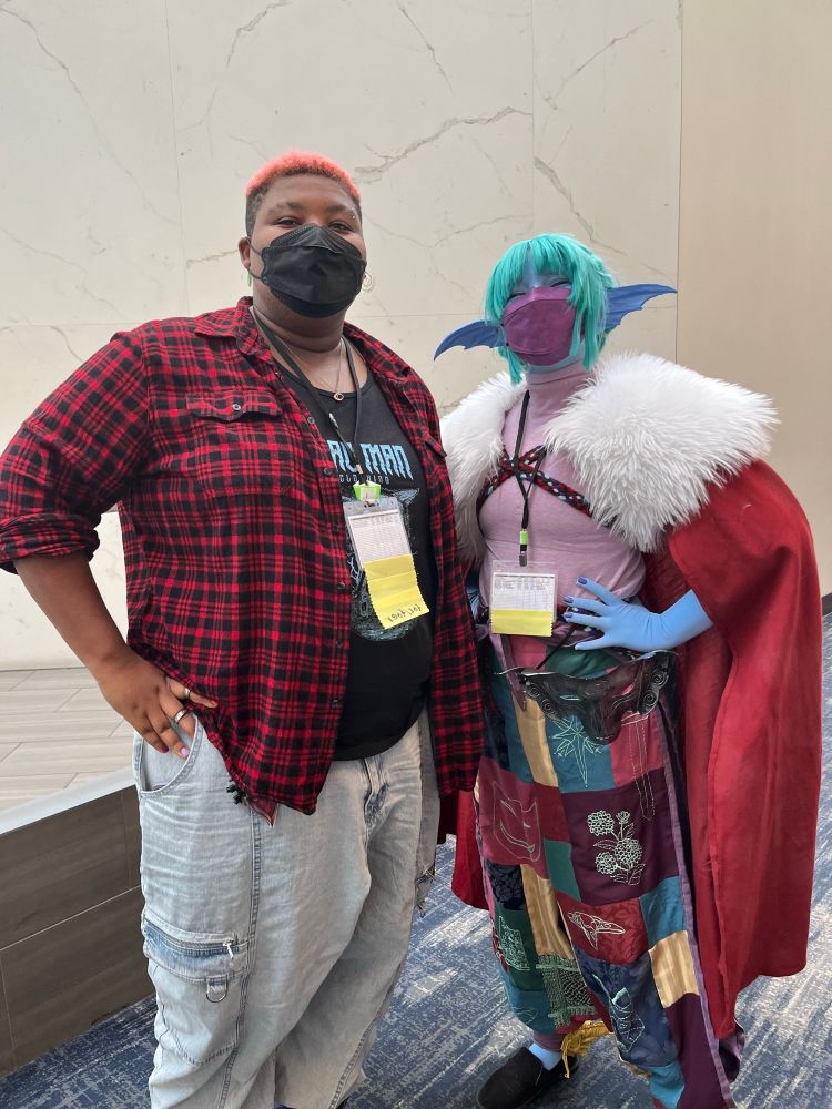 Erika cosplaying Gentle from TransplanarRPG with a purple KN95 and taking a photo with Dare. Dare is a tall Black butch person with short hair dyed pink at the top. She’s wearing a black KN95 mask. Fae are wearing a black and red flannel, light washed baggy jeans, and a black graphic t-shirt. 