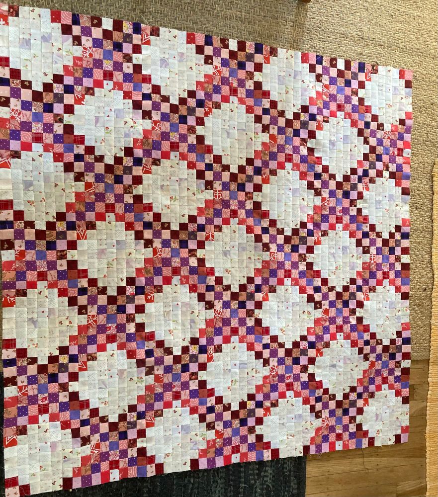 A quilt top (known as a flimsy) made of 1” red, purple, pink, and white squares arranged in a chain link like pattern (triple Irish chain).  The red,purple, and pink squares form diagonal lines across the quilt leaving squares of white.