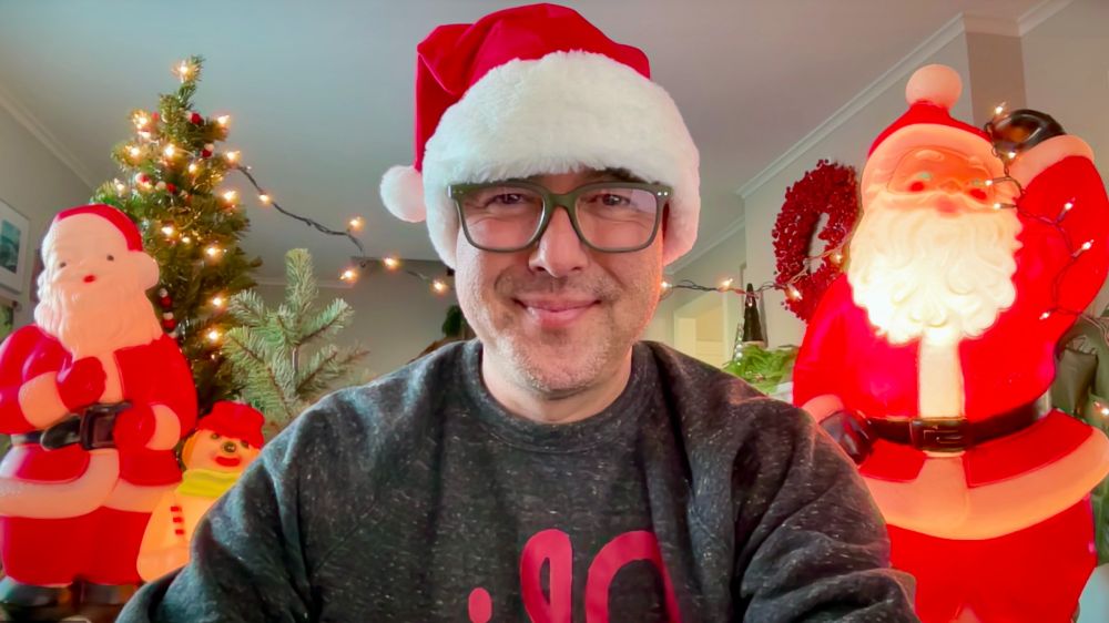 The author wearing a Santa hat and Ohio sweatshirt sitting with blow mold Santas, Christmas trees, and string lights