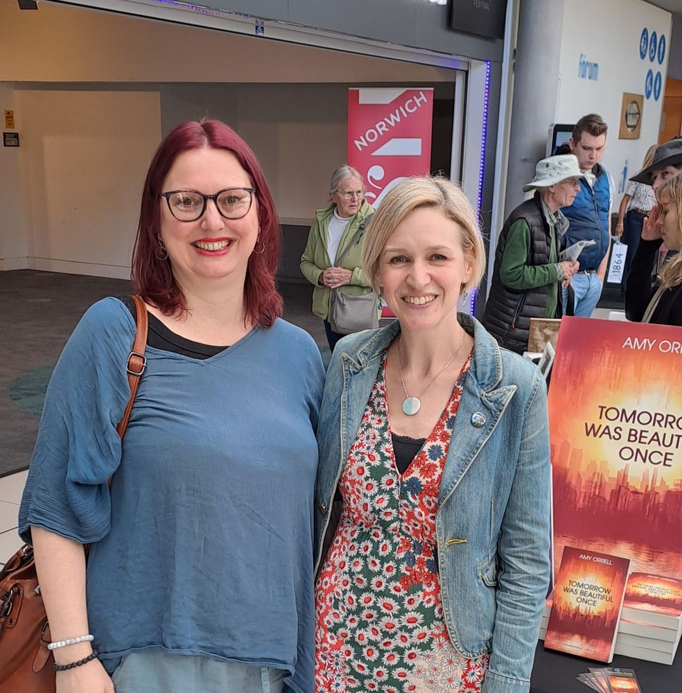 Image is a photo of two women standing together. I am on the left, with red hair, glasses and blue top. Amy is on the right with short blonde hair, a denim jacket and patterned dress. There is a sign behind Amy that is a poster of her book, Tomorrow Was Beautiful Once. In the background is a small group of people.