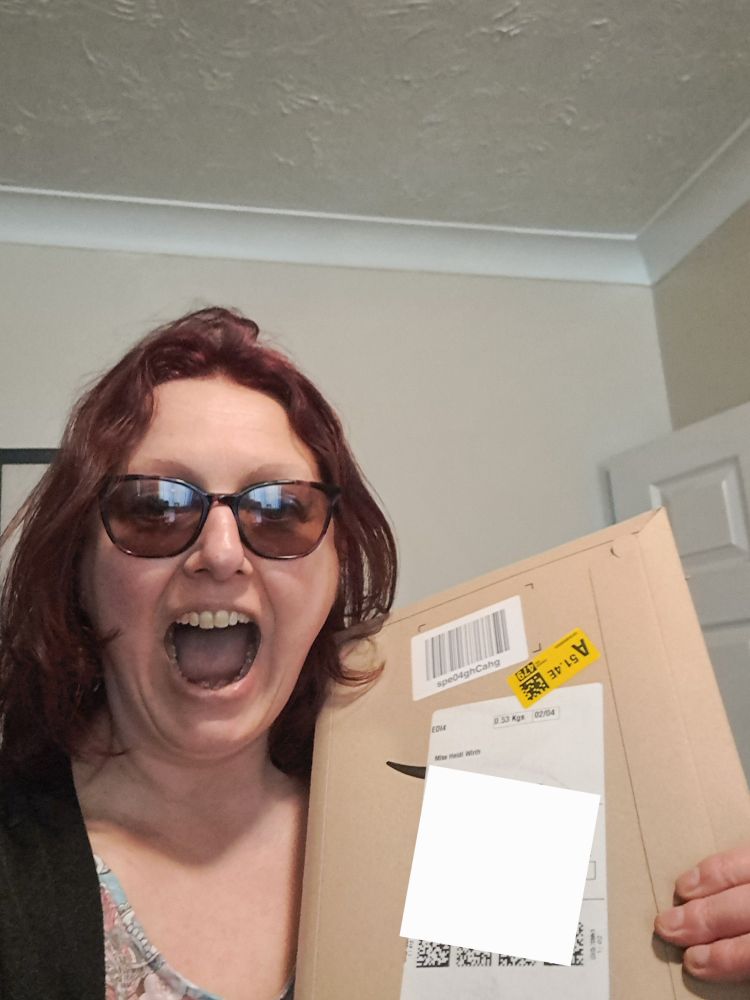 Image is a photo of me, a female, either reddish brown hair, dark glasses, and a mouth open in delighted excitement. I am holding an unopened book size package.