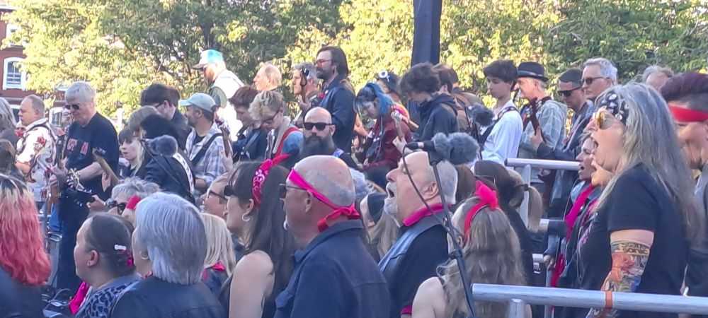 Image is a photo of a crowd of people, some are a choir, and further back are guitarists.