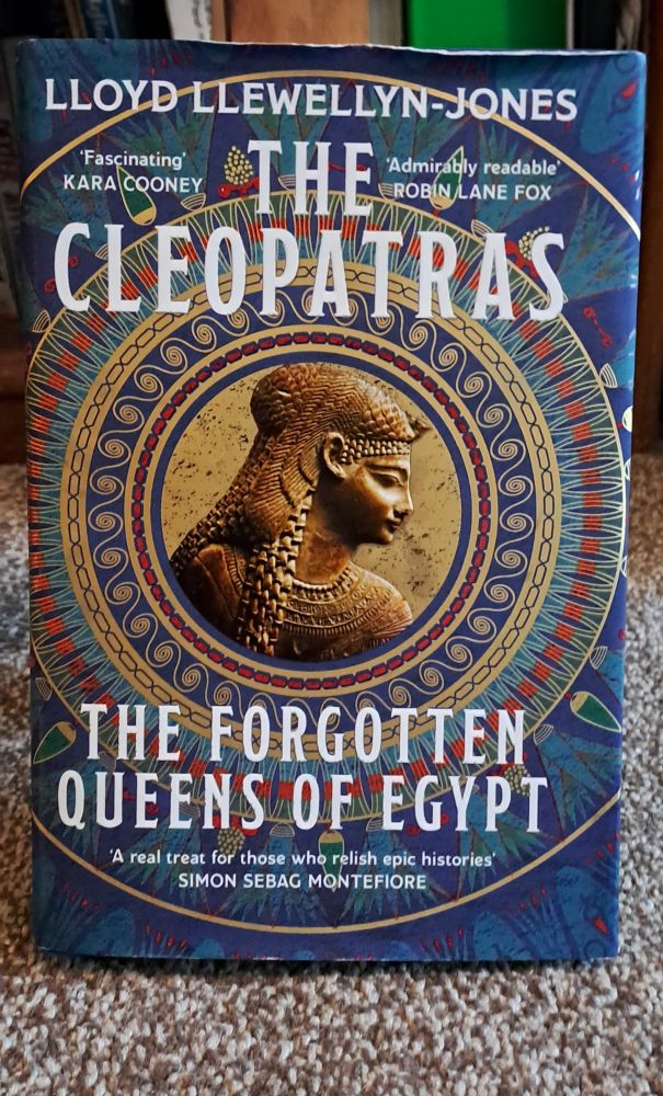 Image is a photo of a stunning book cover. Authors name is at the top: Lloyd Llewellyn-Jones. Title: The Cleopatras: The forgotten Queens of Egypt. The book background is blue, with a patterned circle in the middle of the book, mostly gold, and an image of cleopatra (also gold) in the centre.