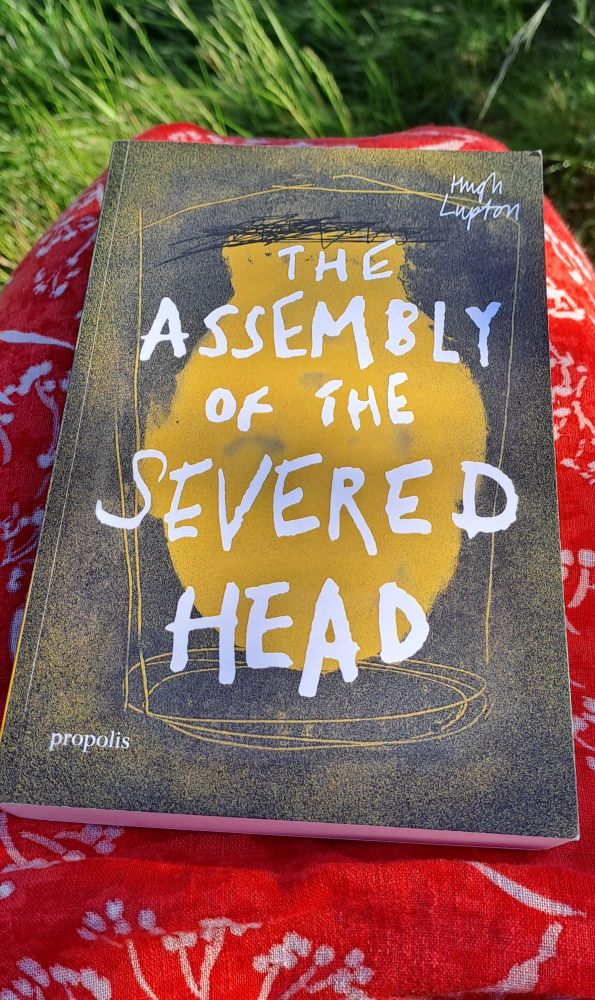 Photo of a book cover, with the title: Assembly of the Severed Head.
The book has a dark background, and an orange vase shape in the centre. 