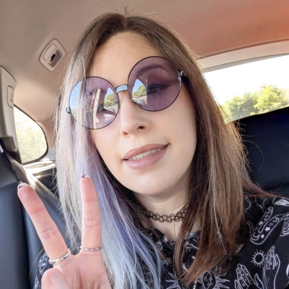 Car selfie - IRL FeliKitty - smiling and doing the victory sign. Wearing round glasses with purple lenses, black shirt with mythical/mystical designs, black choker. 