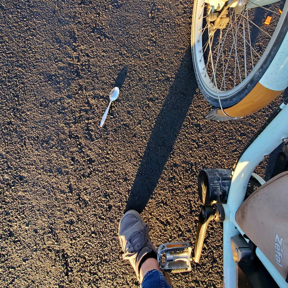 A photo from my point of view astride a bike looking down at the ground. On the asphalt is a shiny metal spoon.