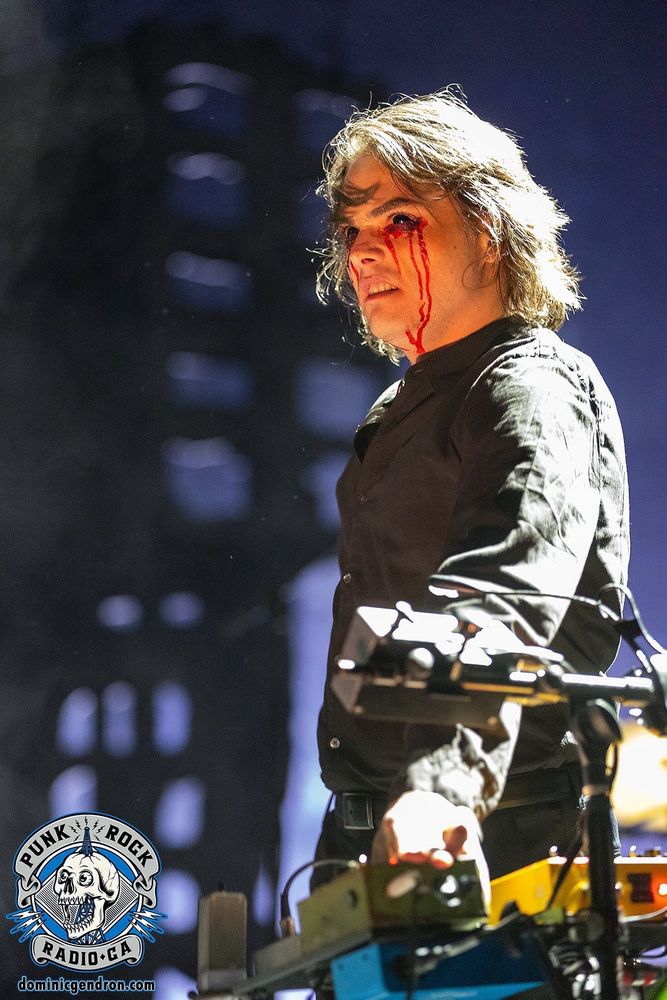 Gerard Way playing with MCR in 2022, dressed in a back button up and bleeding from his eyes. He's also wearing black contacts and looks like he's growling, his hand is on one of his effect pedals. 