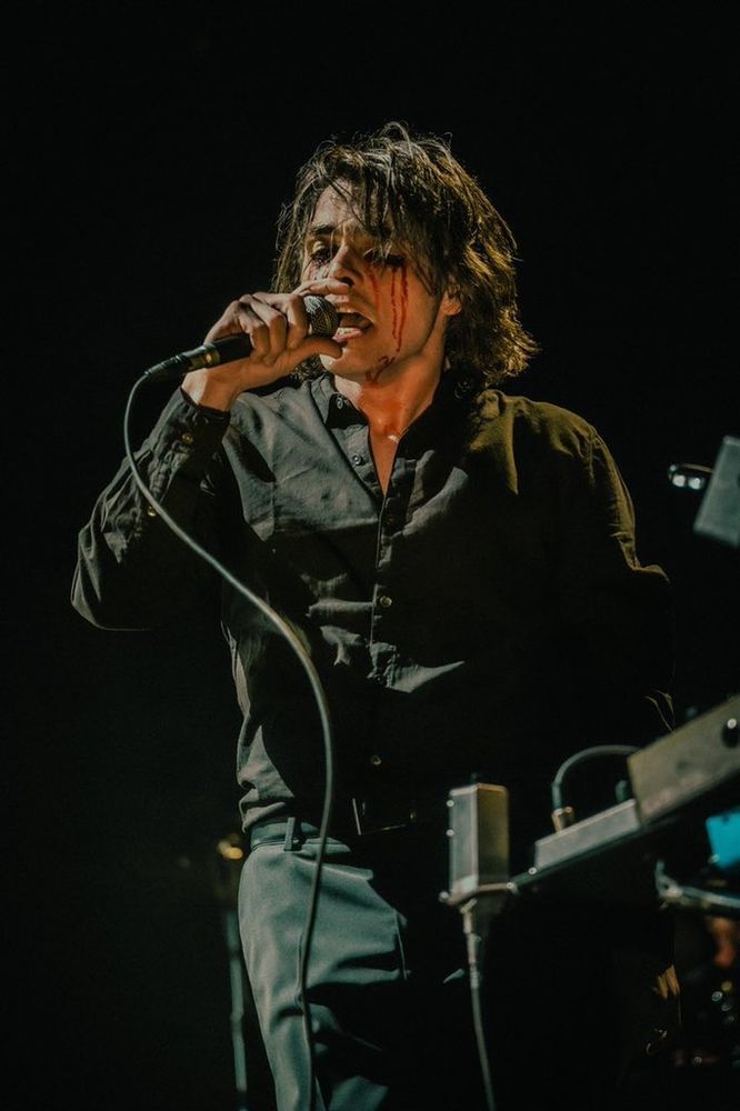 Gerard Way playing with MCR in 2022, dressed in a back button up and bleeding from his eyes. He's also wearing black contacts and singing into the microphone. 