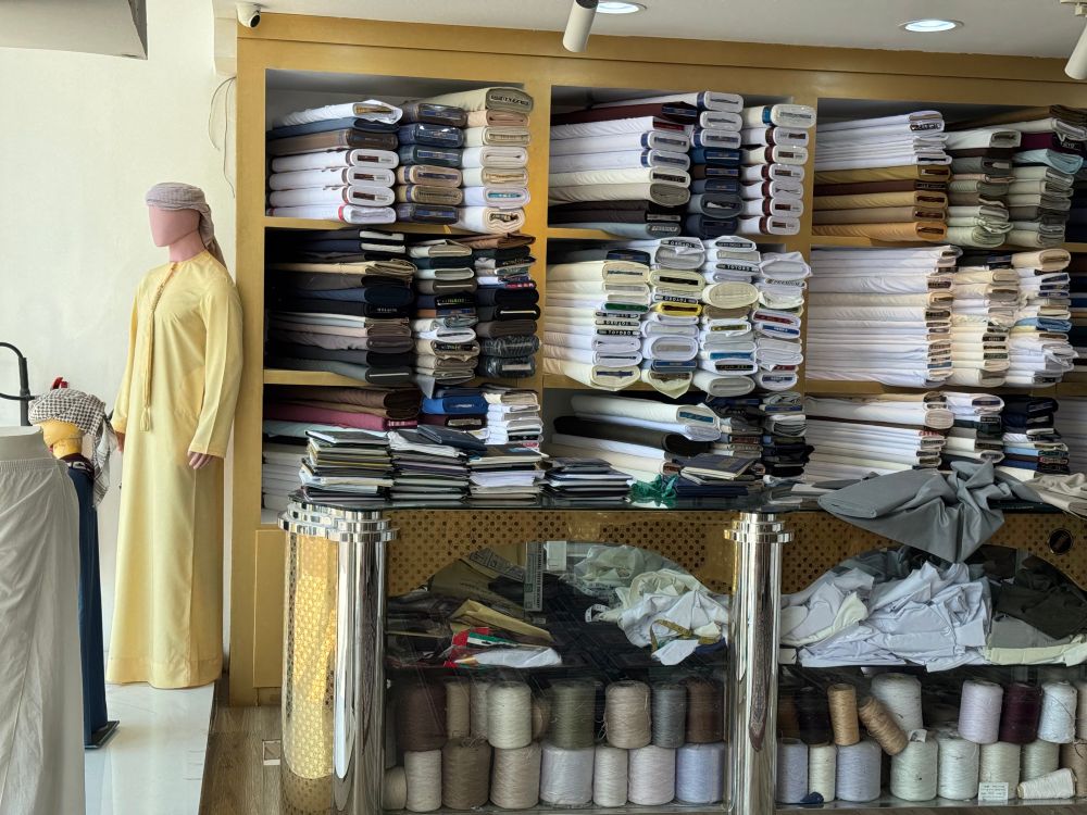 Inside a traditional tailor’s shop, shelves are stacked with neatly folded rolls of fabric in white, beige, and dark shades. A mannequin dressed in a yellow kandura and headscarf stands to the left. Below the counter are spools of thread and folded cloth, suggesting custom garment work.