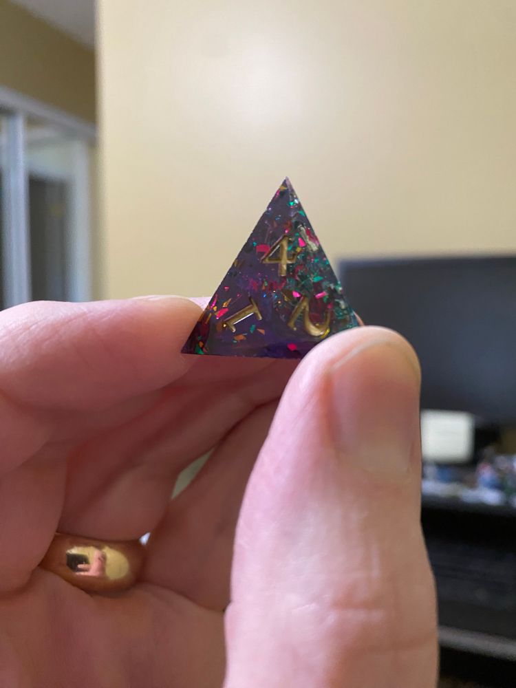 A four sided die in purple and teal with reflective inclusions, numerals painted gold