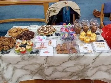 A table covered with a grey marbled effect cloth holds plates of apple crumble traybake, 24 cookies (triple chocolate and chocolate and marshmallow), Millionaire's Shortbread, sweetie bags, gingerbread teddies with little hand-iced faces and scarves, Christmas Sprinkle and Lemon cupcakes, chocolate and caramel cake pots, watercolour Christmas cards and Scrabble style lettered Christmas cards decorated with hand-drawn fairy lights.