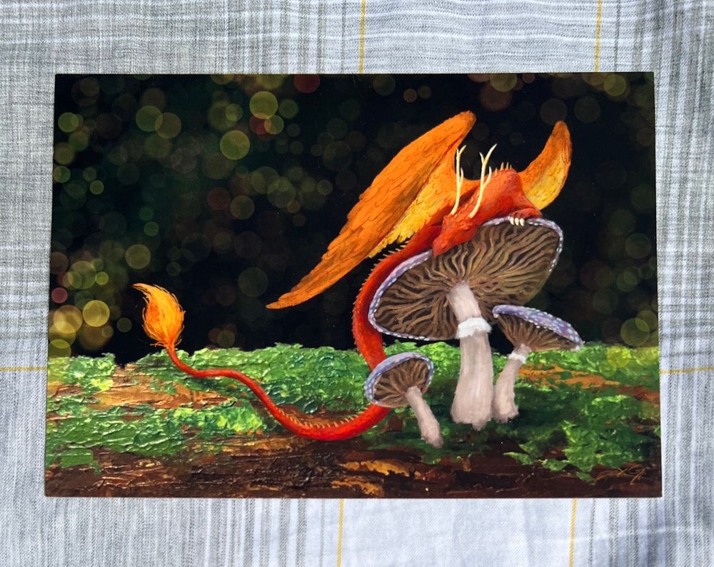 A print of a mixed media piece I did. A little orange dragon snoozes on top of a purple mushroom growing out of a mossy log. The dragon has soft-looking wings and tail tuft, and thin deerlike (branching) horns.

It is mostly a digital painting, but the log is actually a photo of an acrylic painting! The reason is I was working on an all-acrylic version of this piece, but I was so worried about messing up the dragon that I wanted to experiment in digital first. Obviously I ended up liking the digital version a great deal, so I finished it and mailed them out to my Patreon print club! Now I sell them on etsy and at conventions