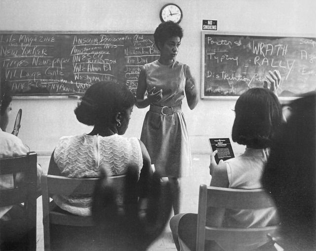 june jordan stands in center commanding a classroom full of eager questions. behind her there is a chalk board full of curiosities that can barely be made out in the black and white photo. 