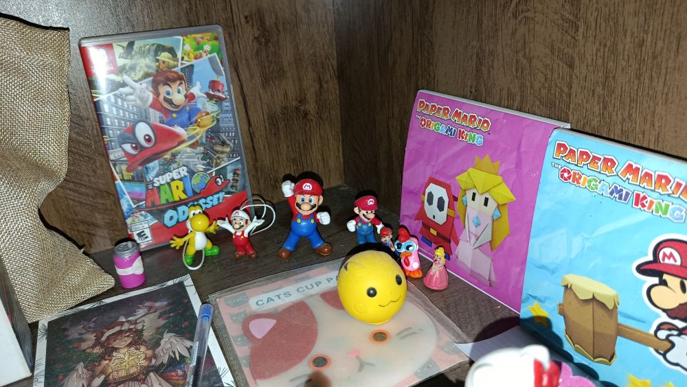 A drawing, super Mario Odyssey game box, a tiny pink can, a tiny yellow yoshi, a tiny fire Mario, a slightly bigger Mario with first up, another tiny Mario, yet another tiny Mario, a tiny princess peach, Pikachu ball, a cat cup stand, a big sticker set with princess peach and a shy guy, another big sticker set of paper Mario