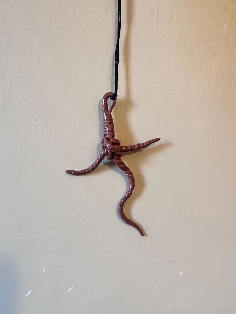 the worm cross - it hangs against a pale eggshell wall on a black cord knotted around the curved tip of the primary worm. one worm makes the stake of the cross and another worm makes the arms. two smaller worms wrap around the center of the cross in knots. the worms in the cross are an ambiguous shade of brownish pink/pinkish brown that swirl around themselves and are glazed in a clear gloss.