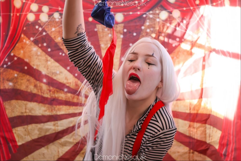 A photo of me, a white haired clown girl, with my tongue out holding a handkerchief dangling in front. I'm in front of a circus background. I'm wearing a black and white stripped shirt with red suspenders.