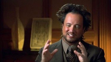 Giorgio from Ancient Aliens wearing a dark grayish suit and hitting his classic pose (his hands up in a this big gesture).

He is sitting in a tasteful, dimly lit room. The sparse lighting is warm and inviting. An old looking stone tablet and long vase add to the vertical-ness of everything in the frame. Even then, Mr.  Tsoukalos hair is still the tallest. 