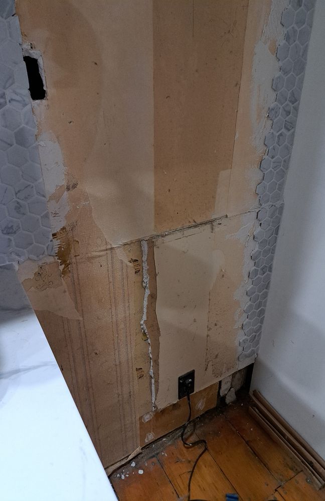 Photo of a wall with patches of tile, broken drywall, plywood, 3 types of old wallpaper, multiple holes, dust, an electrical socket, and the wall doesn't go all the way down to the floor. The plug in the socket seems to have tiling grout on it, suggesting it was left plugged in while work was done on the wall. The floor has an odd misalignment at one point, too.