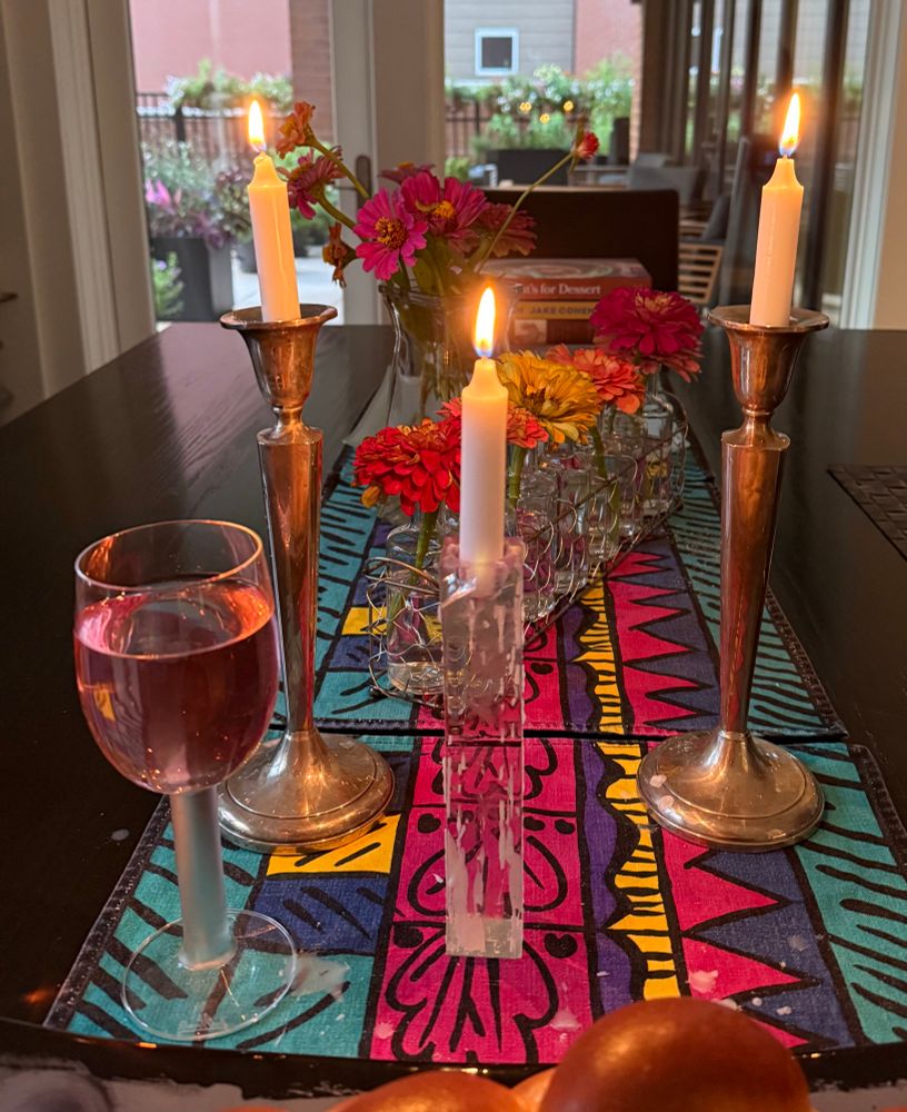 A Shabbat table in front of a window at night. A row of bud vases each with a different color zinnia in front of a tall vase of zinnias; a tall pair of candles; a third one for hope and the hostages; a filled wine glass; partial view of a challah. 
