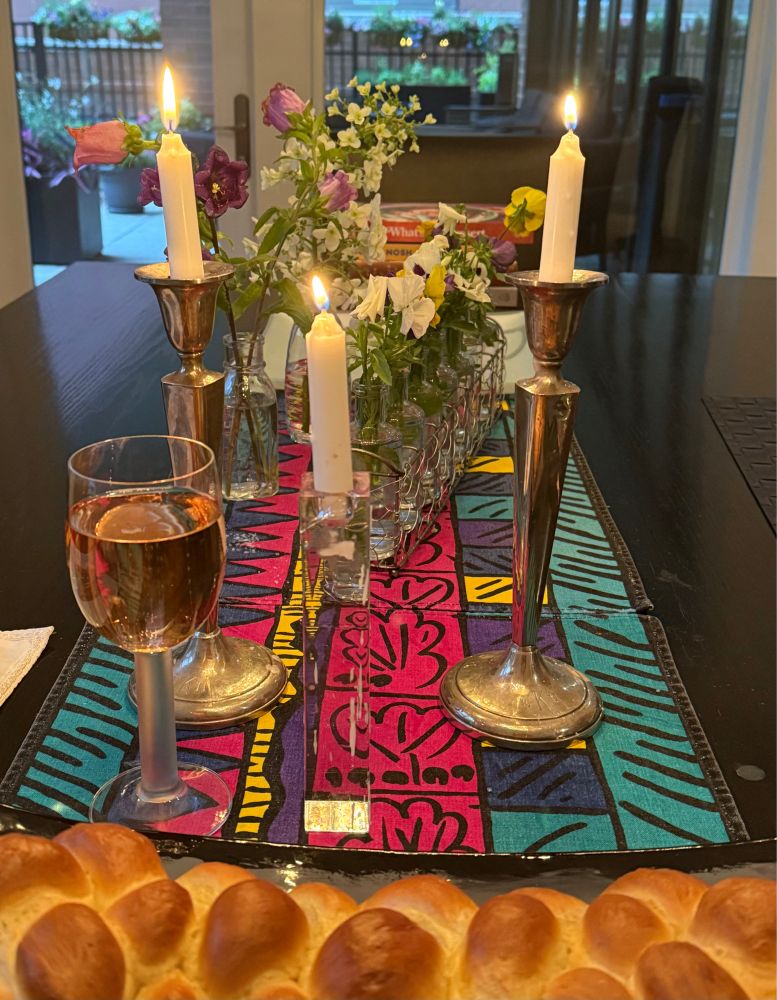 A Shabbat table in front of a window at twilight. Vases of pansies and other garden flowers; a tall pair of candles; a third one for hope and the hostages; a filled wine glass; a challah. 