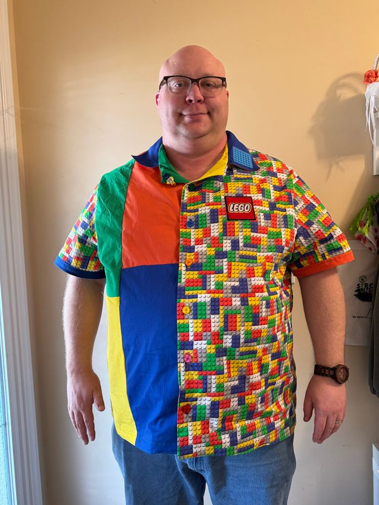 A bald white man wearing a LEGO-themed shirt. Half the shirt is multicolored with LEGO studs on it, and the other half is color blocked in green, orange, blue, and yellow. There is a LEGO patch on the left side of the shirt, a minifigure sewn into the shirt’s right facing, and a sew-on LEGO patch on the left collar.