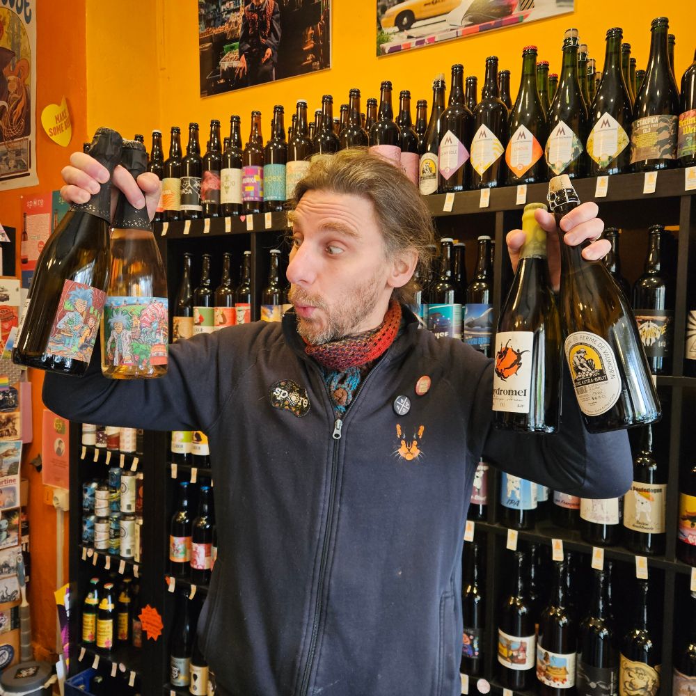 Pierre devant les étagères de la boutique tenant 4 bouteilles : 2 vins pétillants, 1 hydromel et 1 cidre.