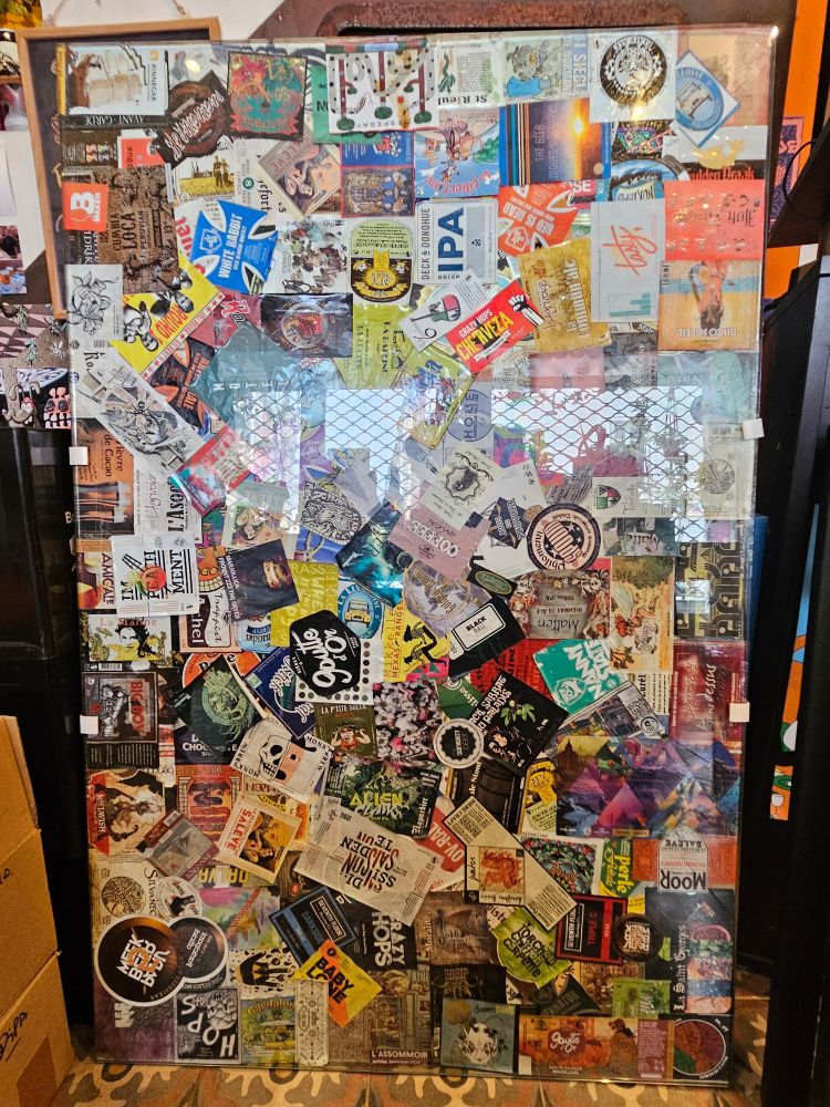 Vitre en verre devant une planche d'aggloméré sur laquelle sont collées une centaine d'étiquettes de bières.