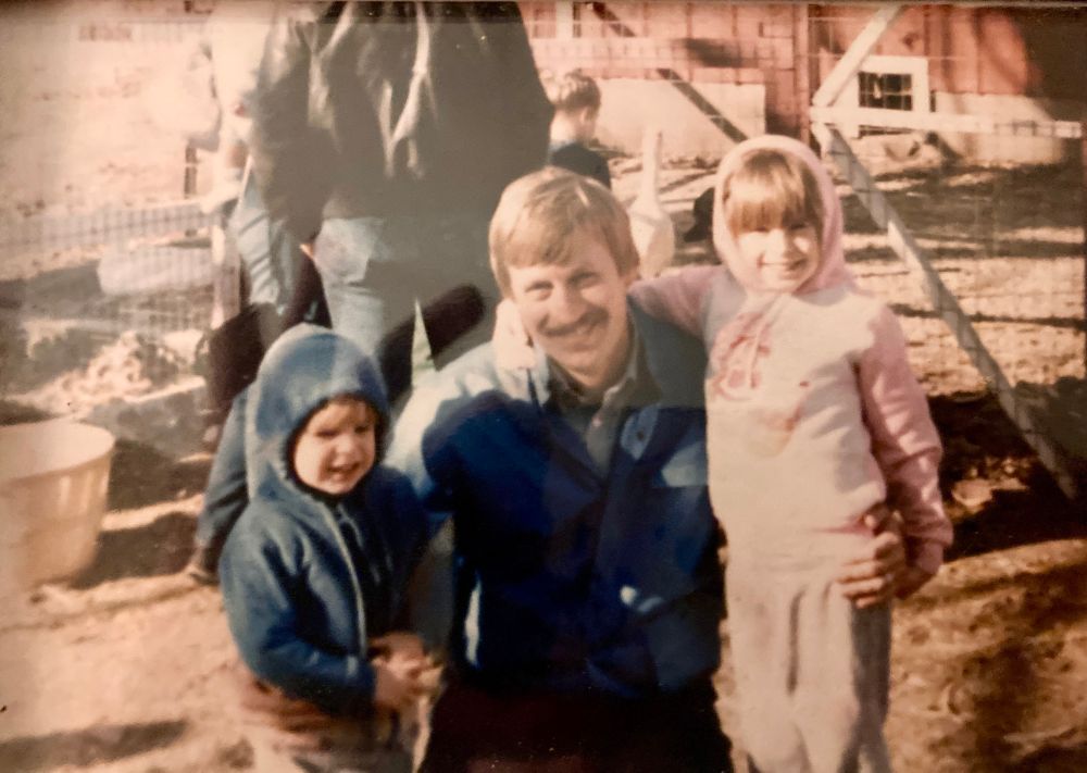 Picture of a 2 year old in a dark blue hoodie with his hands clasped in front of him looking down and off camera to the left of a blond man with a very 70s/80s mustache with his arm around him looking at the camera and smiling with his other arm around a young me wearing a strawberry shortcake hoodie and smiling at the camera with my arm around my dad’s shoulders. 