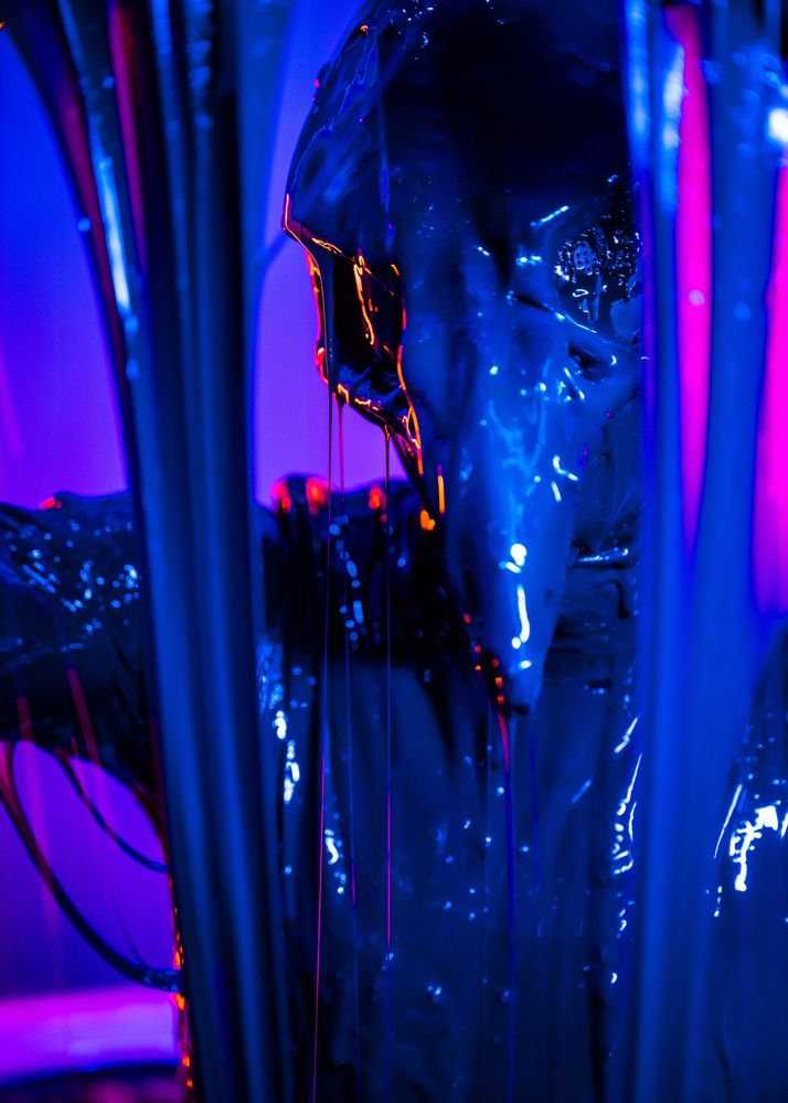 A picture of a rubber bird wearing a latex suit and a crow skull mask basking under a shower of black goo.