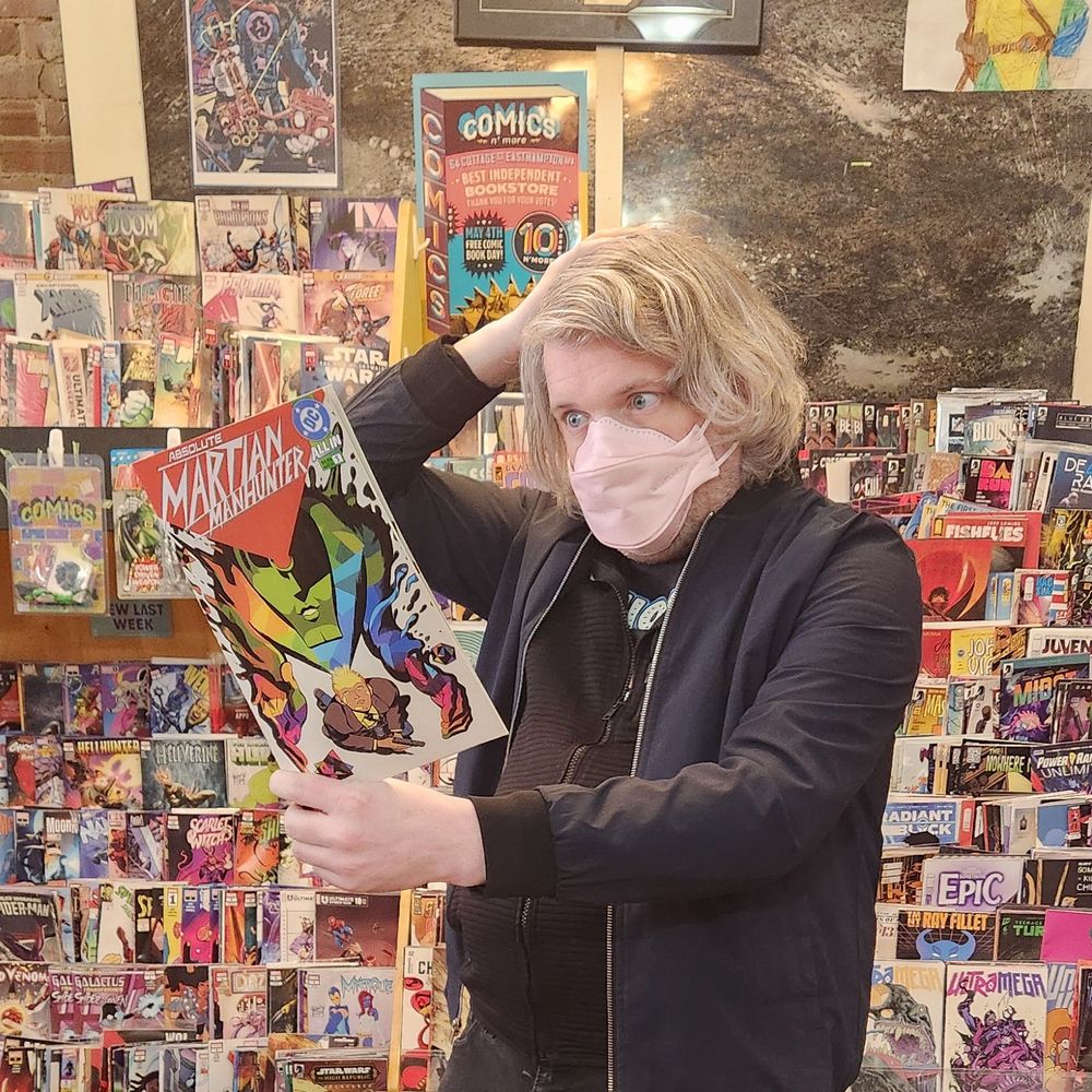 I'm reading the first issue of Absolute Martian Manhunter from DC Comics. I'm wearing a pink kn95 and am surrounded by comics displays in my comics bookstore, Comics N'More.