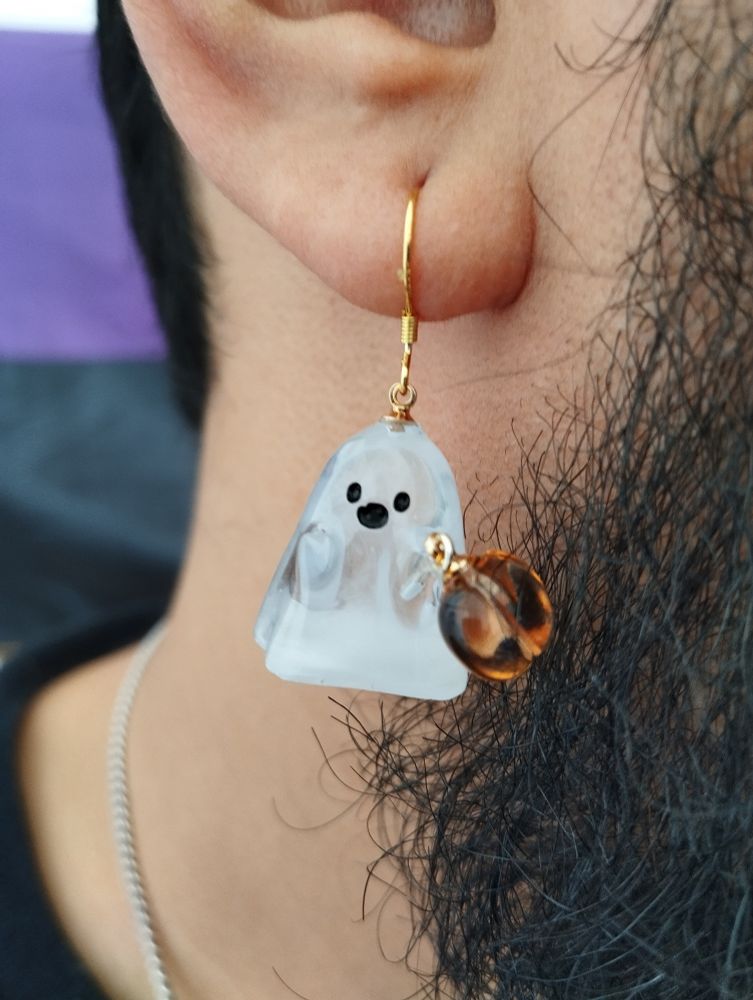 Pendiente con forma de fantasma. El fantasma es blanco pero transparente, y tiene una calabaza en la mano. Los detalles son de acero con acabado dorado.
