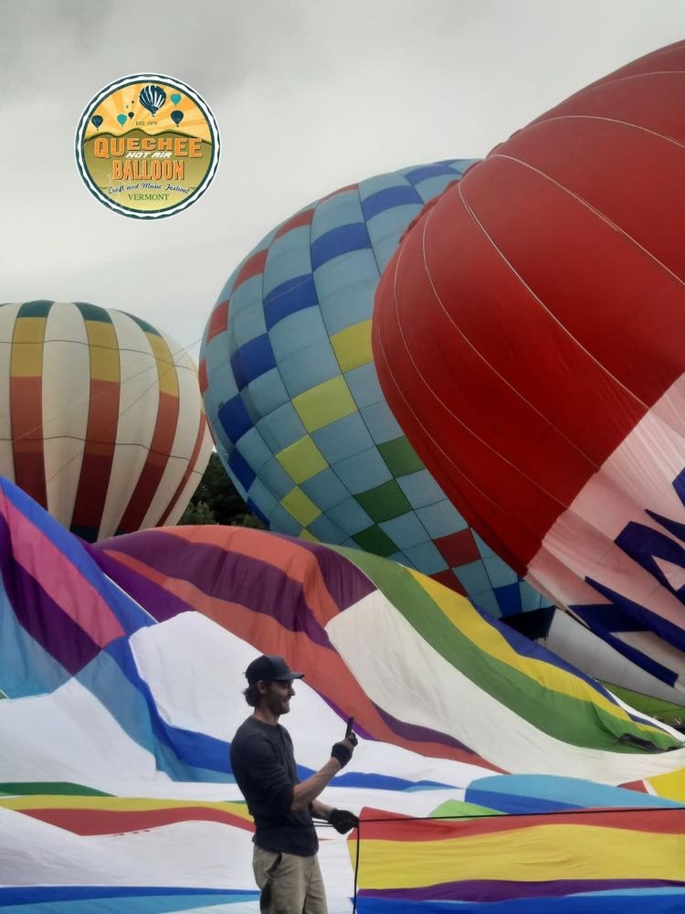 A photo of a man in a ball cap taking a photo in front of four brightly-colored hot air balloons, all of which are in various phases of deflation.
