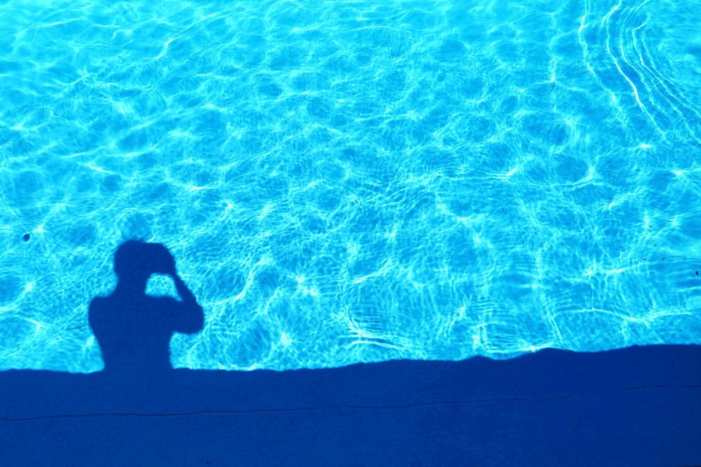 Sur un avant-plan bleu foncé mon ombre en train de photographier une belle piscine bleu clair aux reflets irisés .