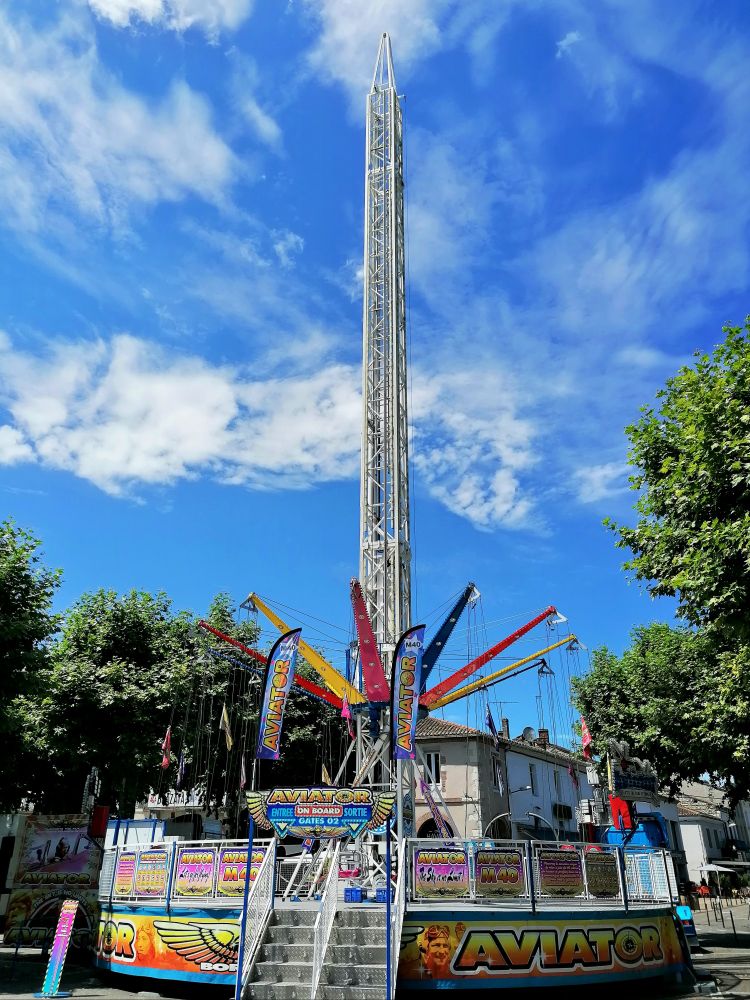 La haute flèche d'une attraction foraine se détache sur le fond d'un superbe ciel bleu. 
À sa base de grands bras métalliques colorés en cercles permettant d'accrocher les téméraires qui voudront  s'envoyer en l'air...
Le manège est festif et très coloré. 
