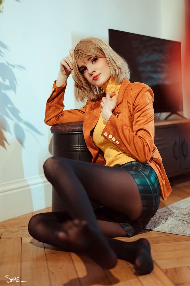 Model is sitting down in costume with black thights, green shirt, yellow top and orange jacket