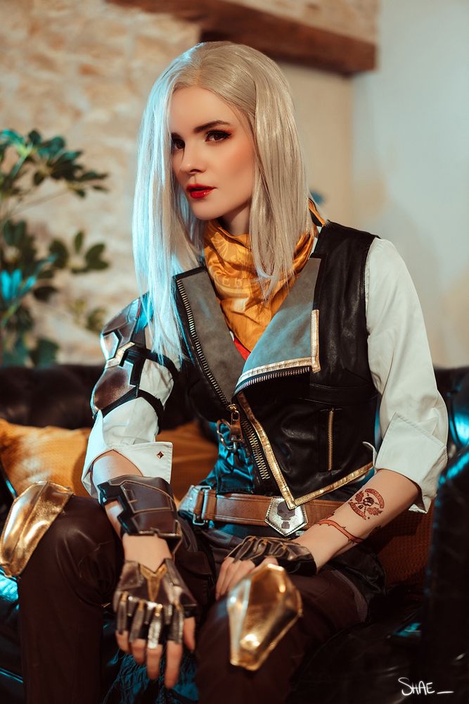 Model is sitting, has a white wig and costume with white shirt, black sleeveless jacket, brown belt, fingerless gloves.