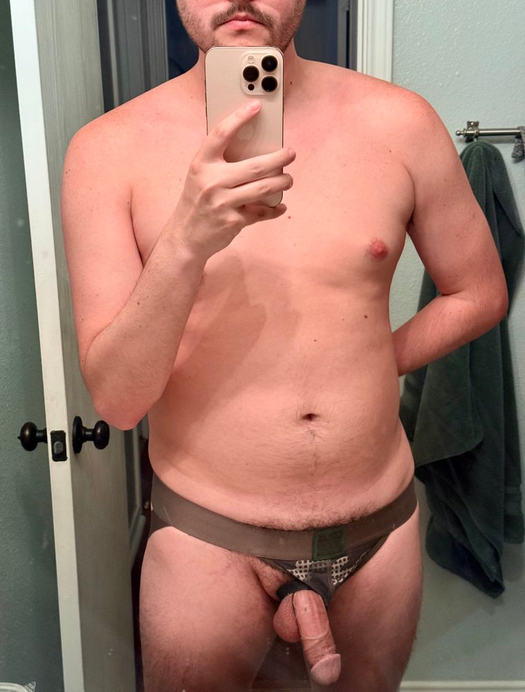 A mirror selfie of Helvetigay cropped from just below his nose to mid thigh, showing exposed chest and a hard cock pulled out the side of a green jockstrap.