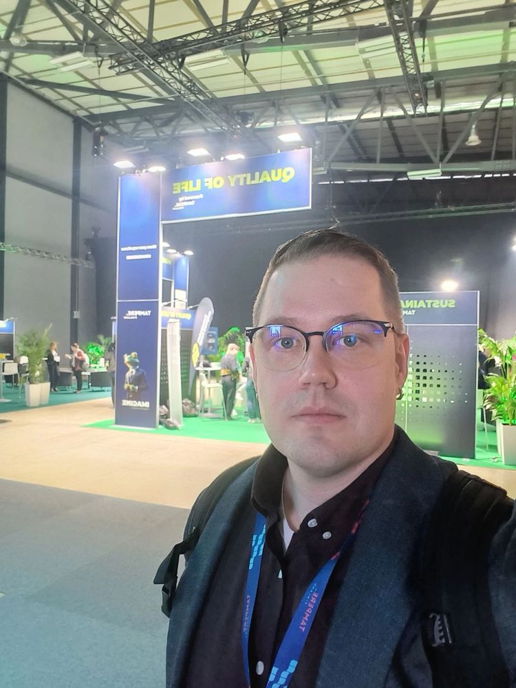 Image shows a conference attendee wearing a lanyard. Behind him is an exhibition for a project.