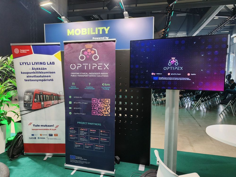 Image shows three banners and a flatscreen television in a conference venue. The first banner is for the Lyyli Living Lab, OptiPEx and Mobility. The television shows a video for the OptiPEx project.