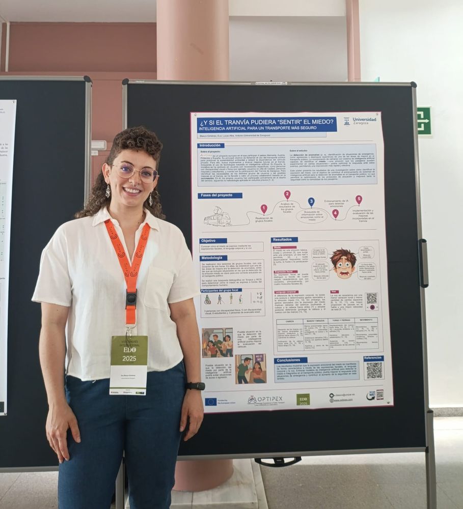 Image shows a participant at a conference wearing a lanyard and badge. The speaker stands beside a poster representing a research project.