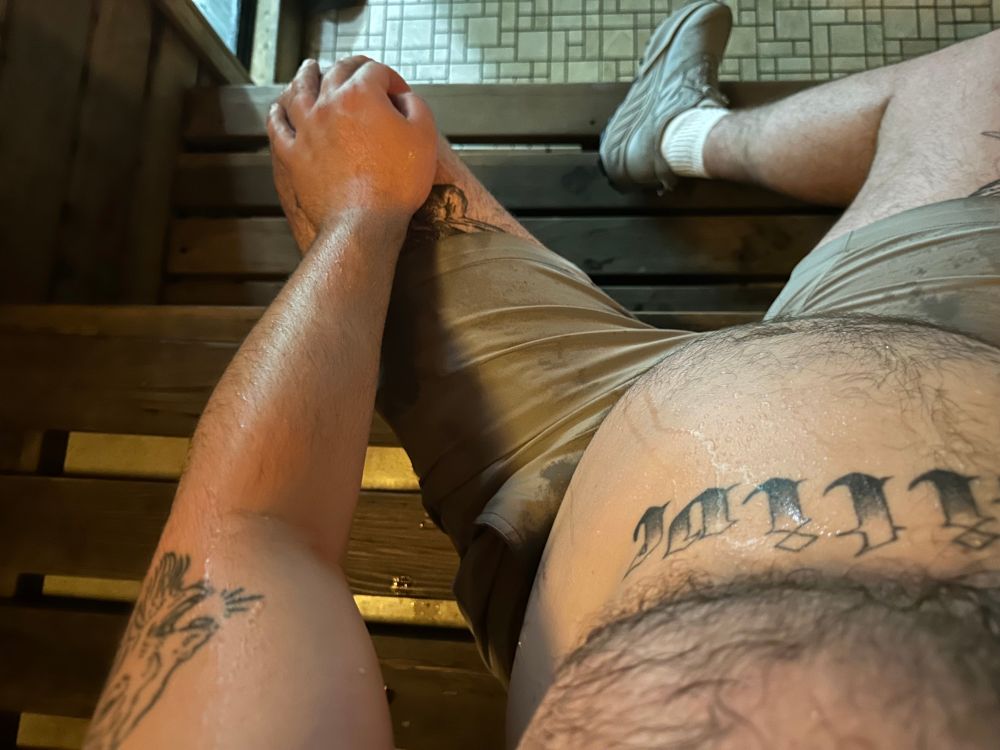 POV shot of me sitting down in a sauna. My tattoos and belly are visible and you can see sweat dripping down my belly and starting to soak my shorts.