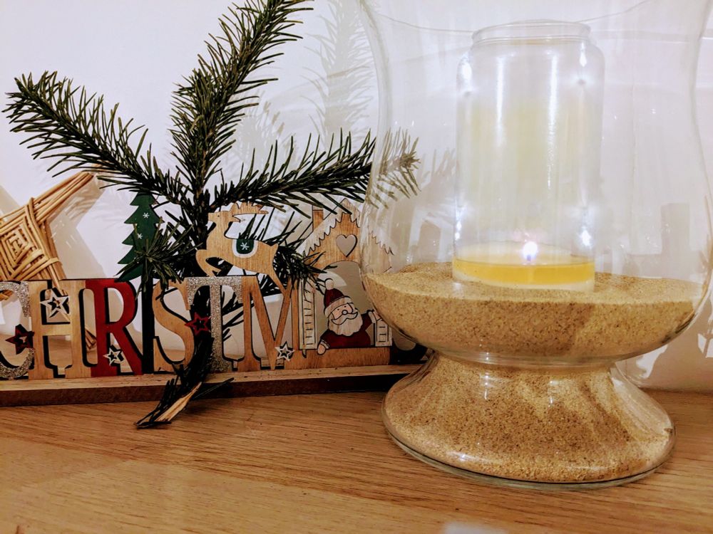 Photo avec à gauche un décor en bois peint marqué ''Christmas'' et à droite un vase en verre contenant du sable et une bougie allumée.