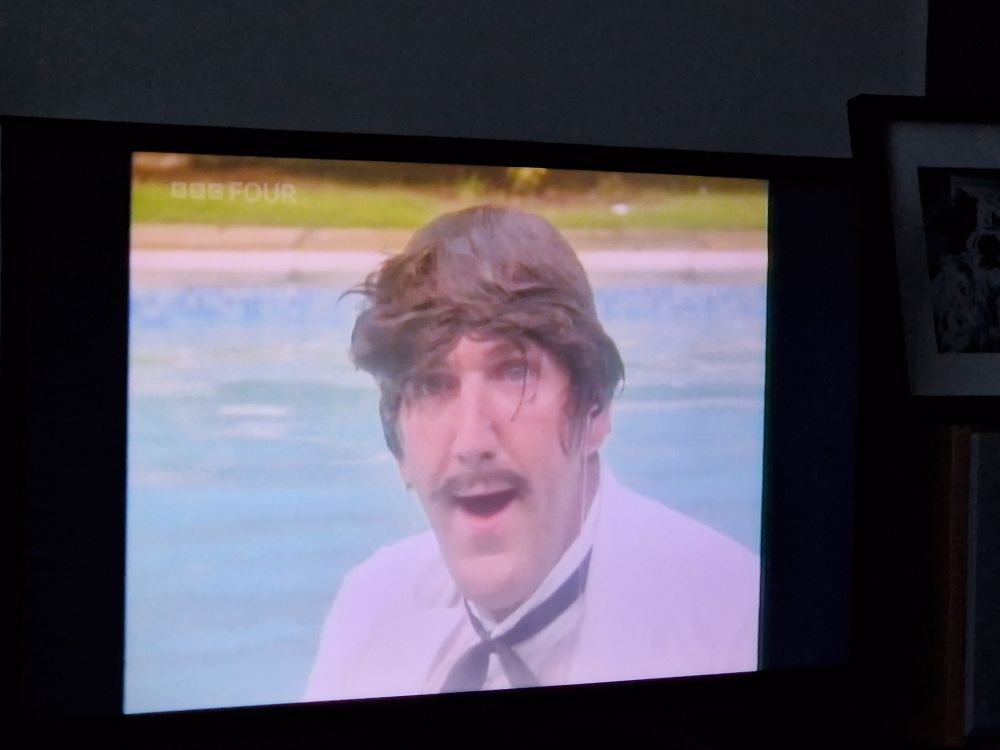Russ Abbott standing in a pool, soaking wet, dressed as a waiter in a bad wig and fake moustache pulling a mugging expression at the camera.  Photo of music video on a tv.