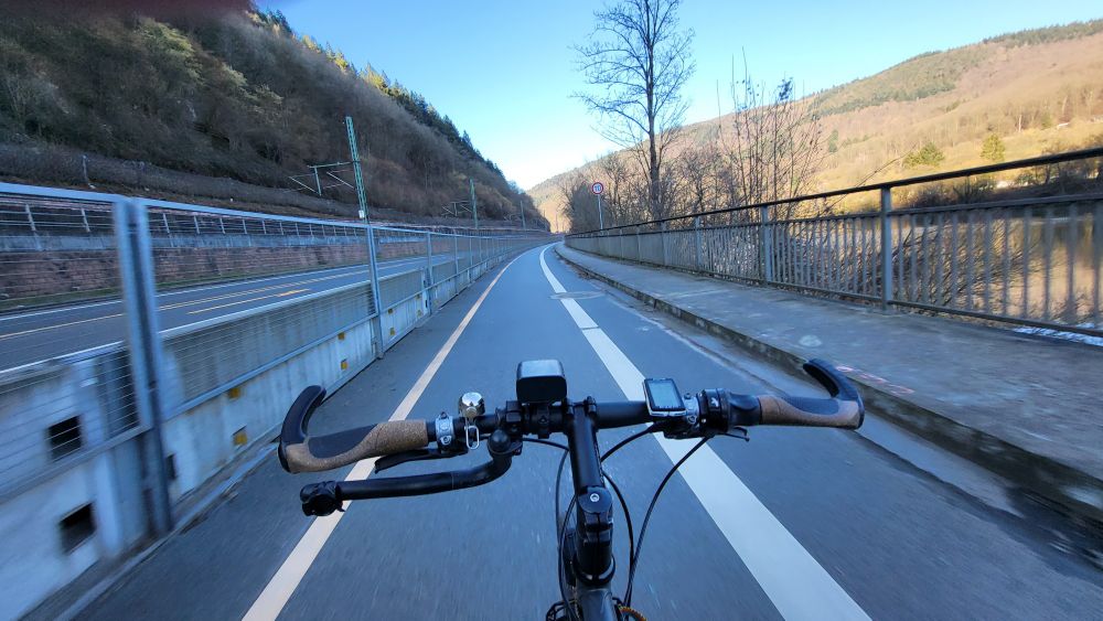 Fahrradlenker aus Sicht des Radfahrers.
Fahrenden auf einer breiten Spur,die durch eine massive Beton-Gitterwand vom Rest der Straße abgetrennt ist. Rechts davon ein Fluss.