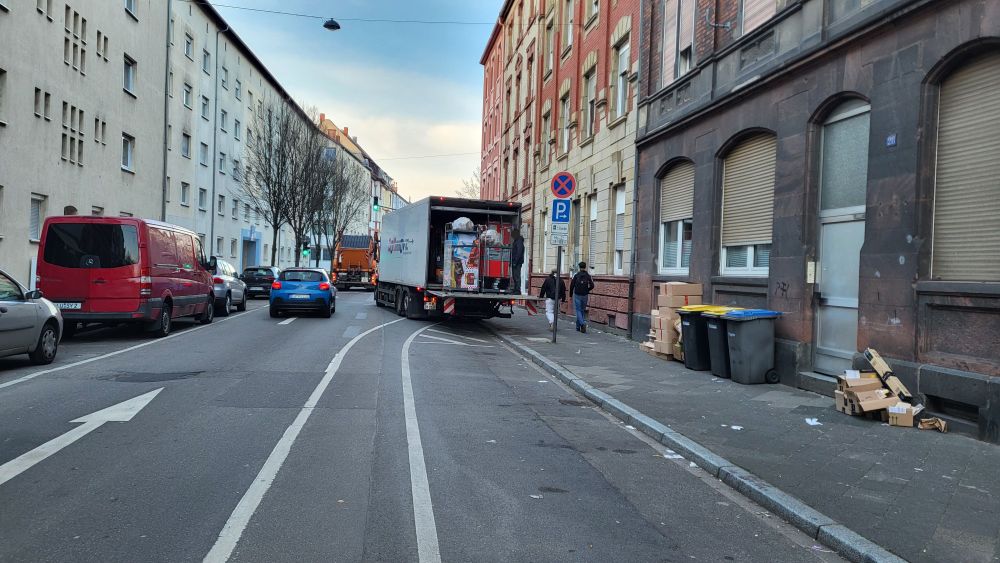 Einbahnstraße. Viel freie Fläche im Vordergrund. Im Hintergrund ein LKW, der schräg auf Fahrbahn und Radweg steht mit geöffneter Ladeluke.