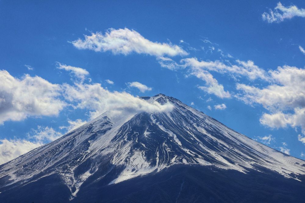Mount Fuji under a bright blue sky with wispy white clouds floating by. The mountain has a sporadic covering of snow.