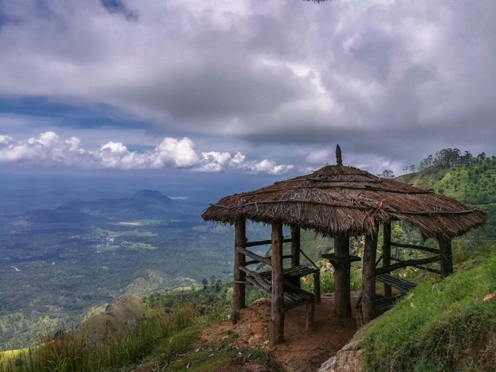An open-walled hut with a thatched roof perched on the edge of a mountain under grey and white clouds overlooking a vast terrain of trees and open land.