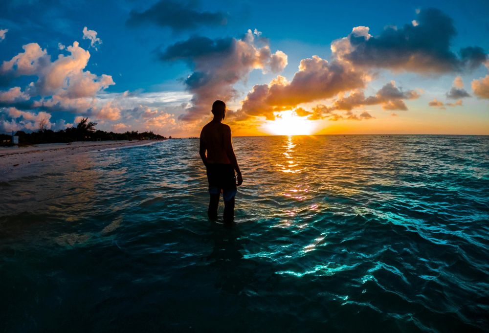 Silhouette of a man standing in shallow aqua-blue water on a beach shoreline, looking at a bright sun setting on the horizon below white fluffy clouds.